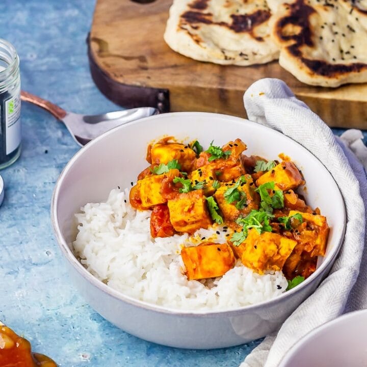 Side angle shot of paneer tikka masala in a grey bowl with naan bread in the background over a blue background