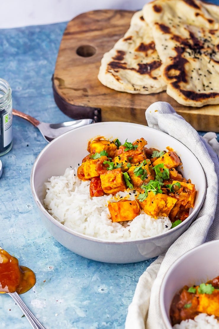 Side angle shot of paneer tikka masala in a grey bowl with naan bread in the background over a blue background