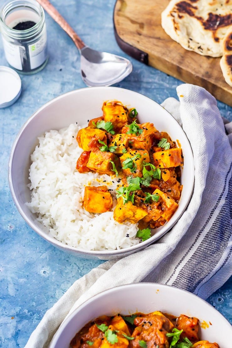 Bowls of paneer tikka masala on a blue background with naan bread and a cloth