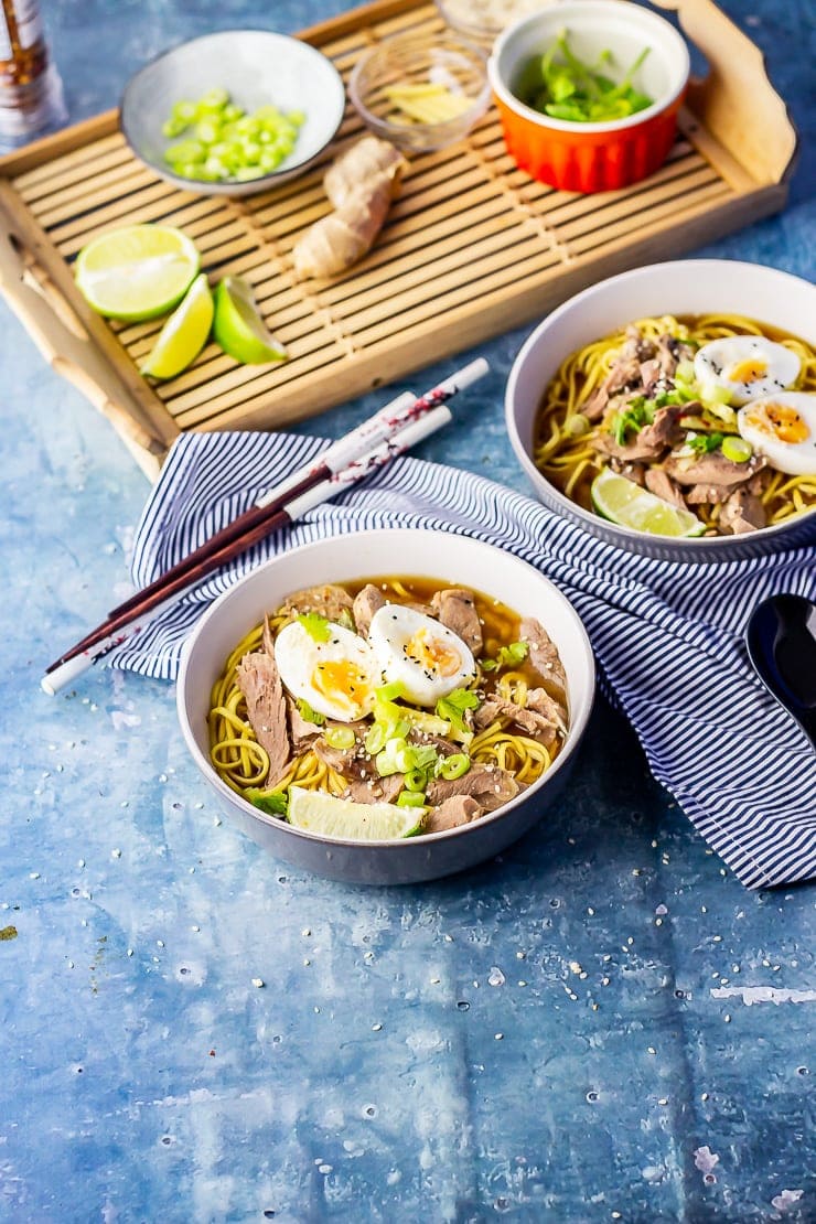 Two bowls of leftover turkey soup with noodles on a blue background with a tray of ingredients