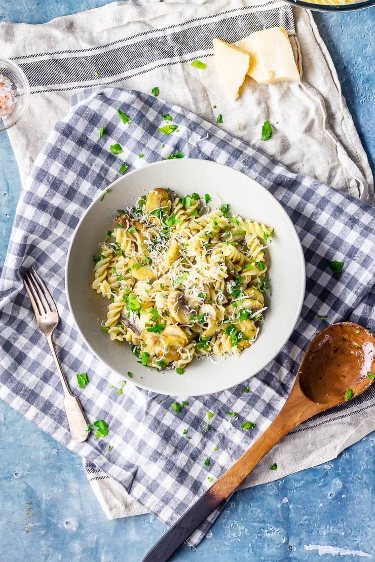 Overhead shot of garlic mushroom pasta on a checked cloth with a wooden spoon and a blue background