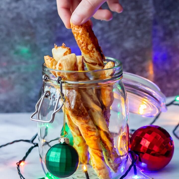 Side on shot of chipotle cheese twists in a glass jar with a hand taking one and Christmas decorations