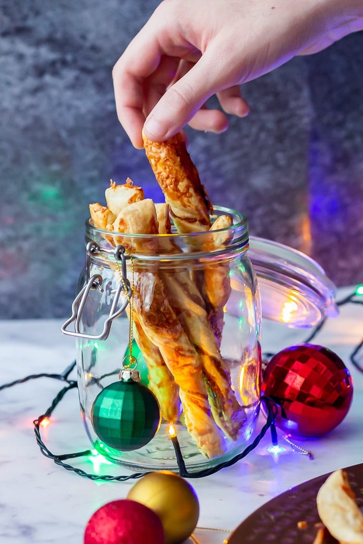 Side on shot of chipotle cheese twists in a glass jar with a hand taking one and Christmas decorations