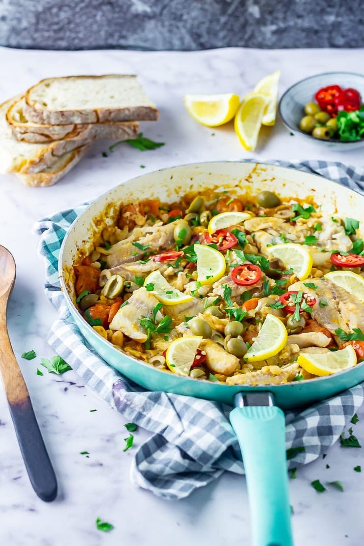 Side angle shot of one pot fish stew in a blue frying pan on a checked cloth with bread in the background