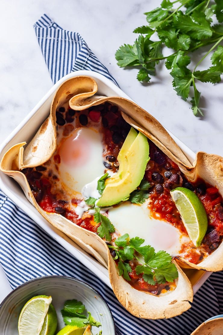 Overhead shot of huevos rancheros baked eggs on a striped cloth with limes and coriander