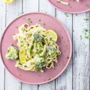 Side angle shot of salmon pasta with broccoli on a pink plate over a white wooden background