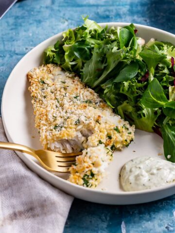 Gold fork cutting into breaded fish with salad in a white bowl