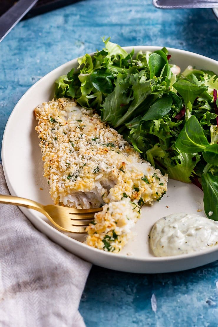 Gold fork cutting into breaded fish with salad in a white bowl