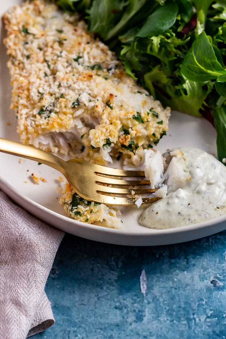 Close up of parmesan breaded fish with a golden fork taking some and dipping it in pesto yoghurt dip