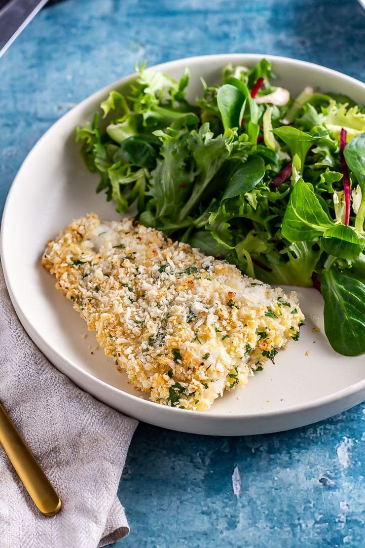 Side on shot of parmesan breaded fish with salad on a white plate over a blue background