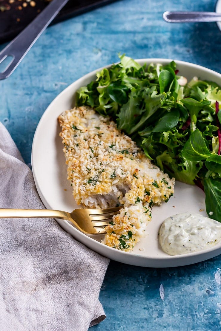 A gold fork cutting into parmesan breaded fish on a white plate with salad and dip