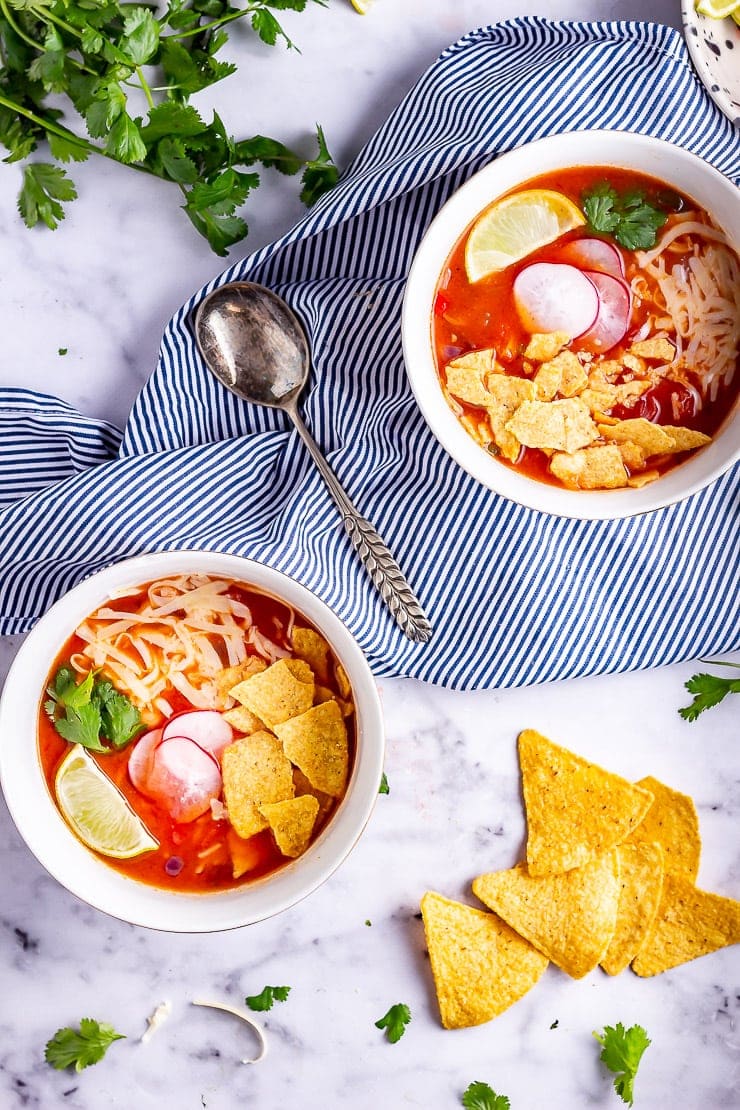 Two bowls of spicy soup on a marble background with a striped cloth and tortilla chips