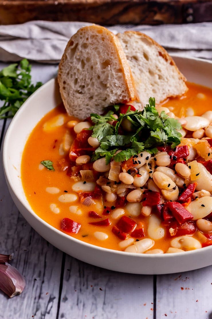 Close up of roasted red pepper soup in a white bowl with bread and herbs