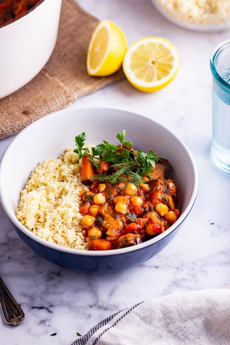 Harissa chickpea stew with couscous and herbs in a blue bowl on a marble background with lemon