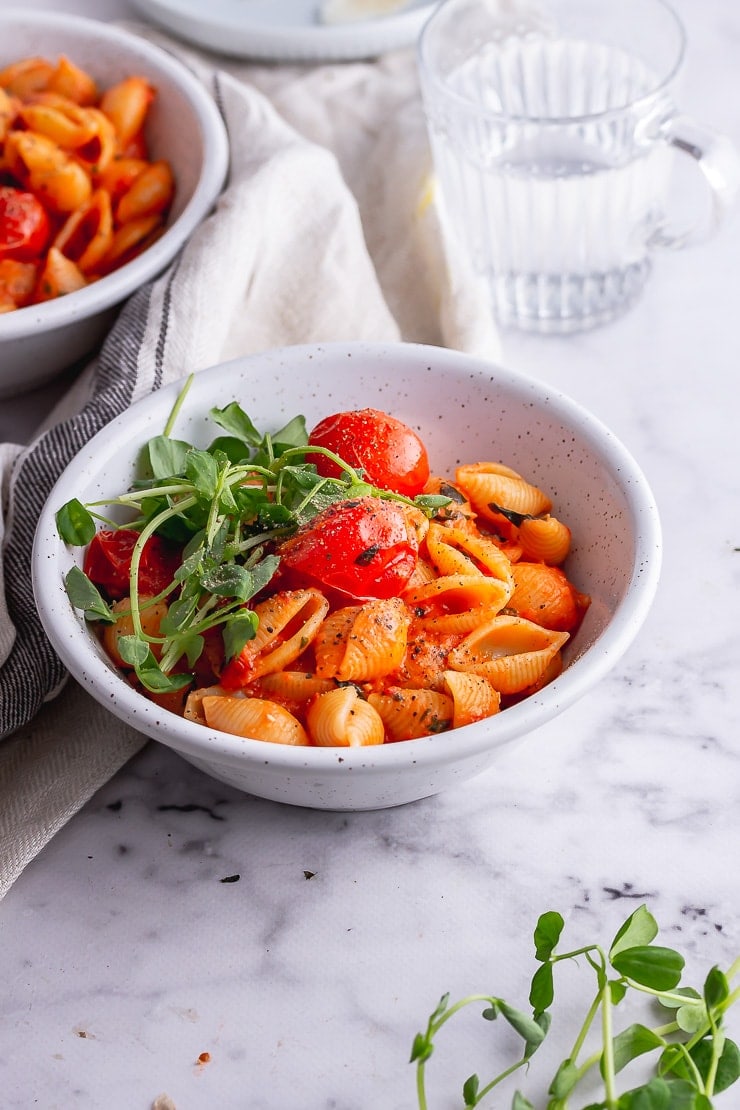 White bowl of tomato pasta with green on a marble background