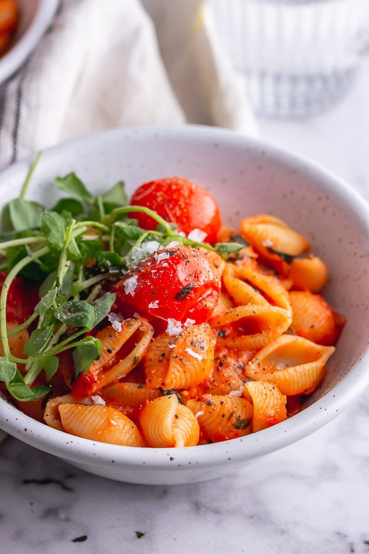 White bowl of tomato pasta with greens on a marble background