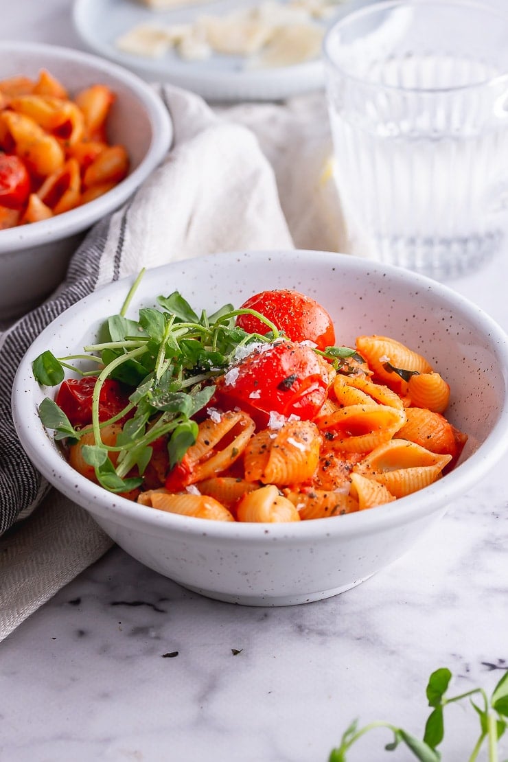 White bowl of tomato pasta with greens on a marble background