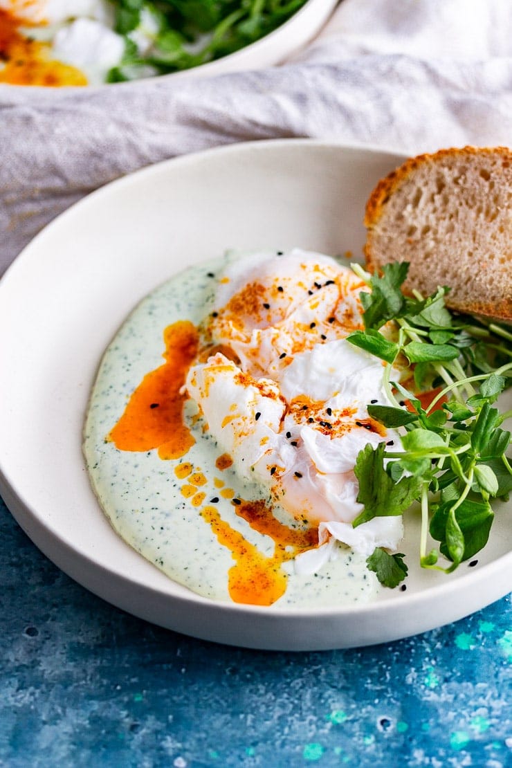 Turkish eggs in a white bowl topped with greens and served with bread all on a blue background
