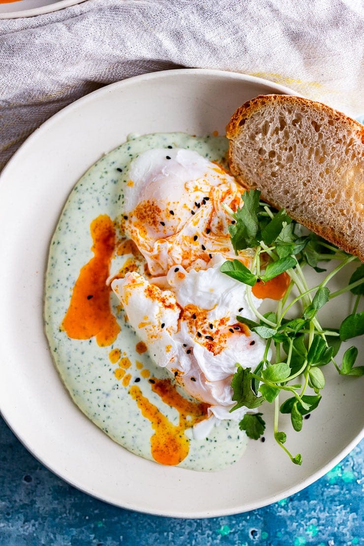 Overhead shot of Turkish eggs in a cream bowl with bread on a blue background