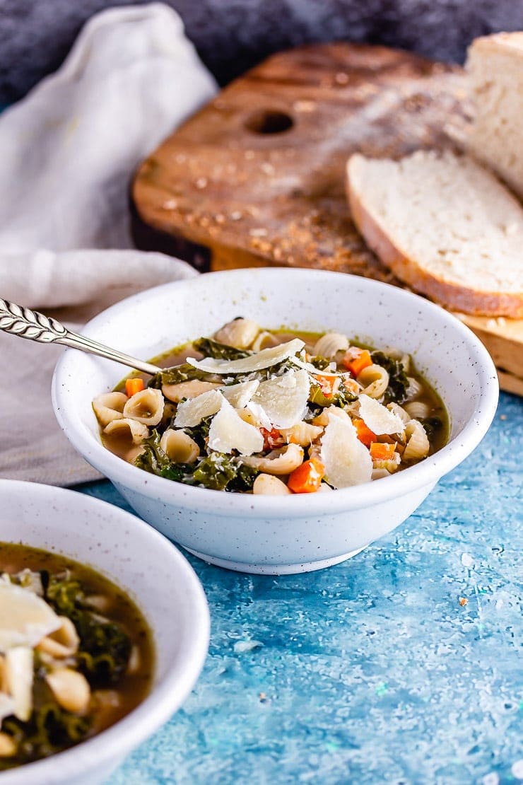 Bowl of veggie pesto pasta soup with bread on a board in the background