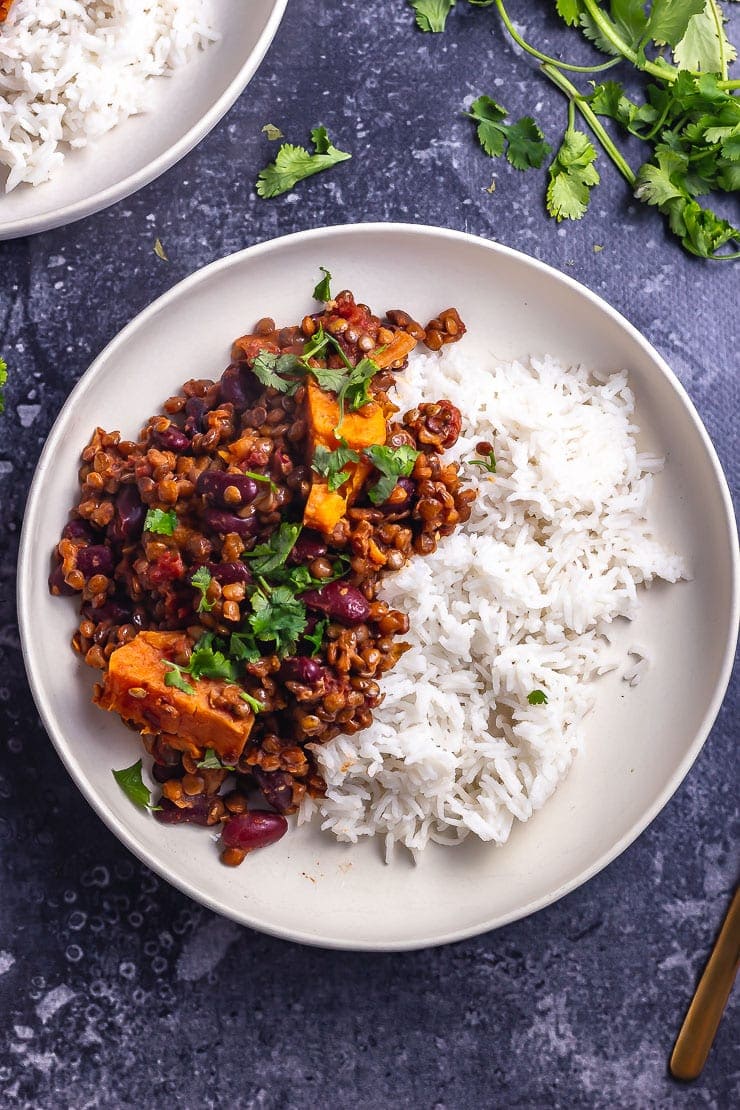 Overhead shot of a white bowl of veggie chilli with rice on a grey background with coriander