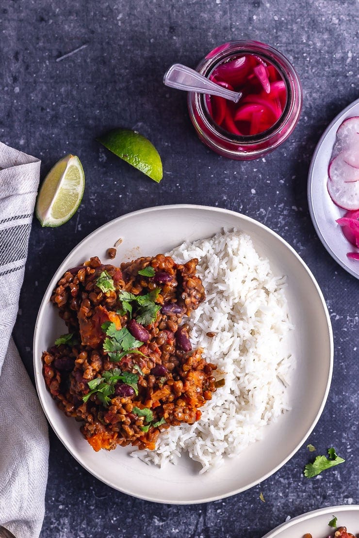 Overhead shot of veggie chilli with rice in a white bowl on a dark background with limes and pickled onions