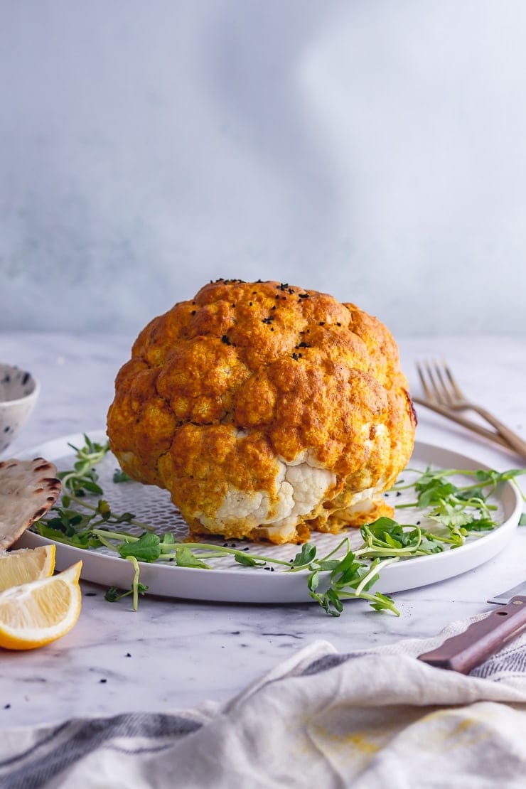 Side on shot of whole roasted cauliflower on a platter with greens and lemon slices