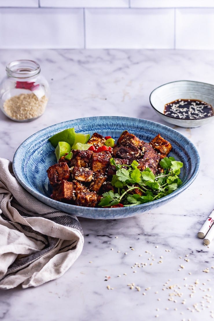 Blue bowl of salt and pepper tofu on a marble surface with soy sauce and sesame seeds