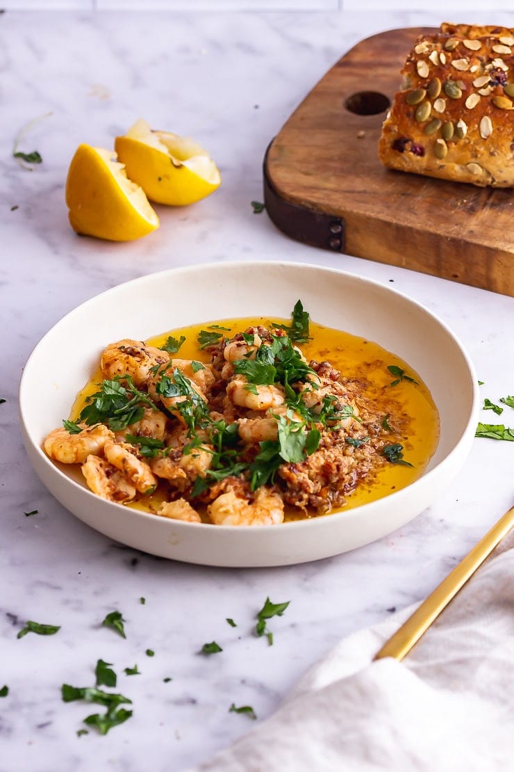 White bowl of chilli prawns with bread and lemon on a marble background