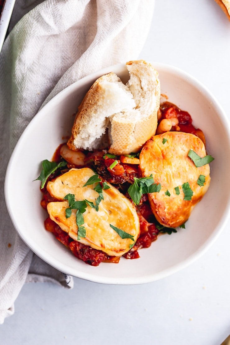 Overhead shot of a white bowl of halloumi, kale & white bean bake on a grey background