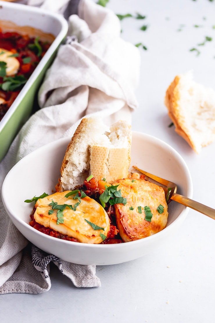 White bowl of halloumi bake with bread on a grey surface