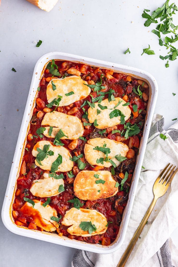 Overhead shot of halloumi, kale & white bean bake on a grey background with bread and herbs