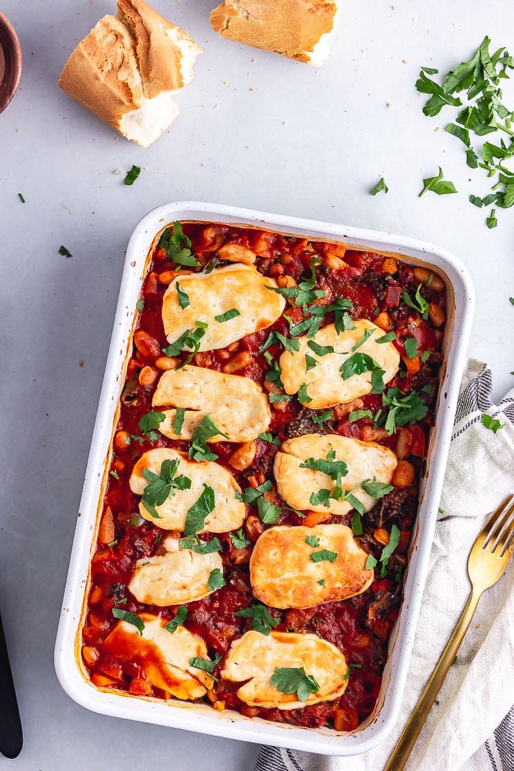 Overhead shot of halloumi bake with beans on a grey background with bread and herbs