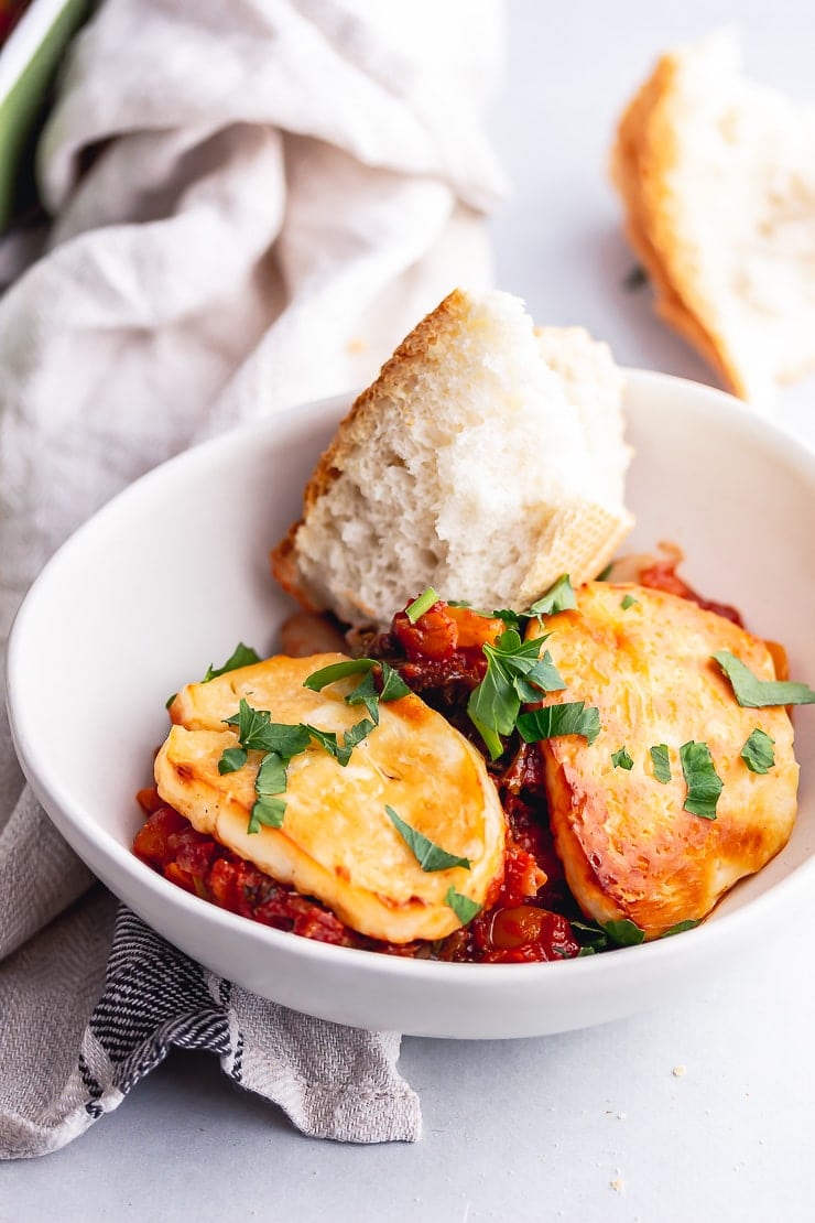 White bowl of halloumi, kale & white bean bake with bread on a grey background