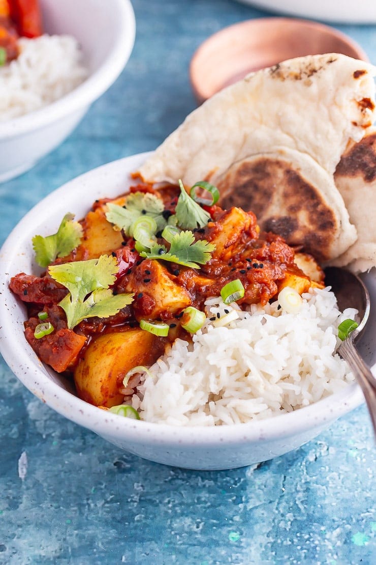 White bowl of halloumi curry with potato with rice and naan on a blue background