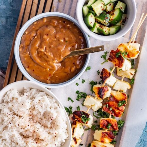 Overhead shot of grilled halloumi skewers with satay sauce, cucumber salad and rice on a wooden tray