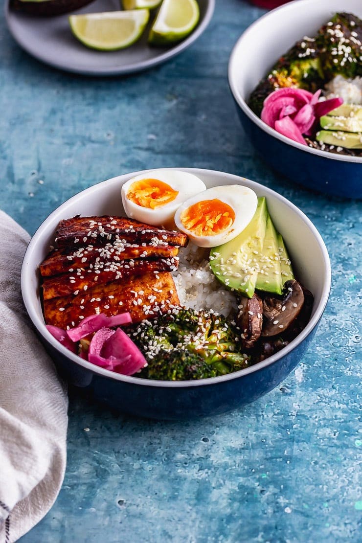 Korean baked tofu in a rice bowl with avocado and egg on a blue surface