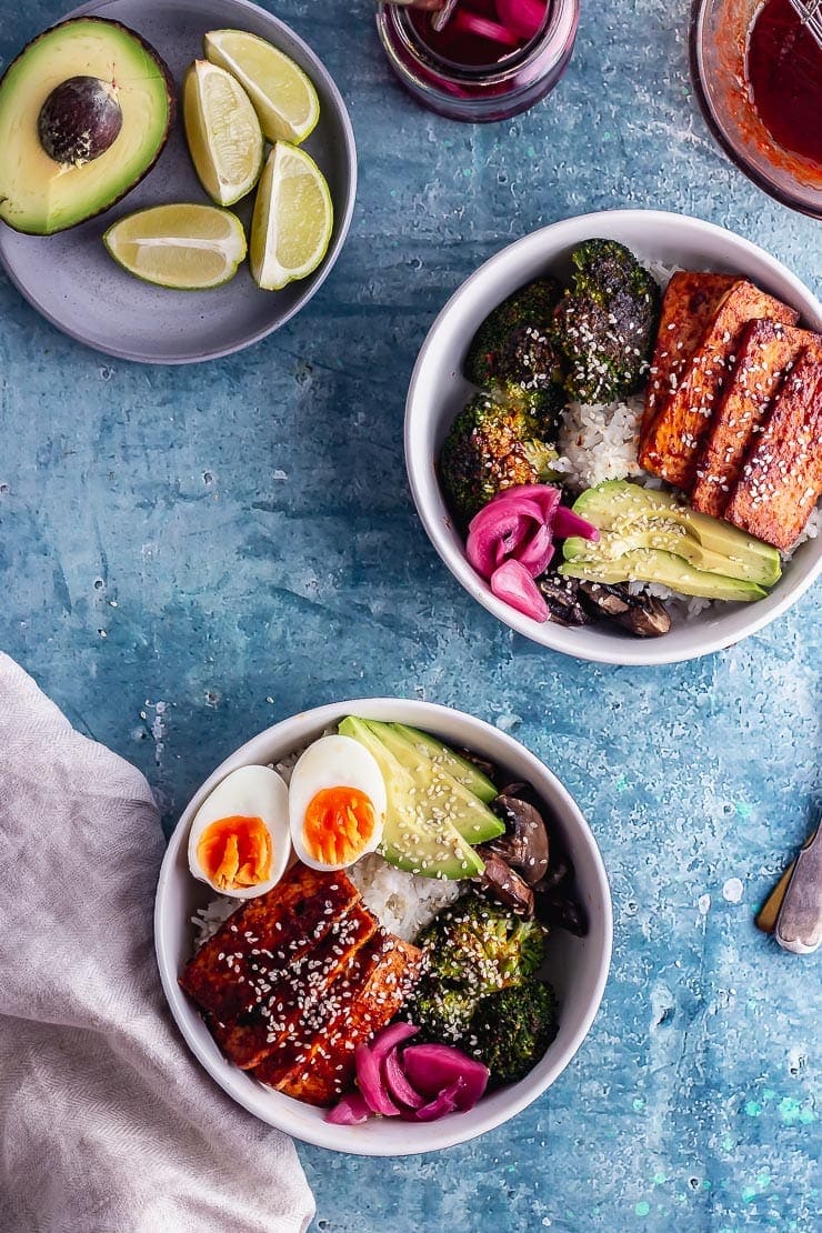 Overhead shot of two bowls of baked tofu bowls with avocado and lime on a blue background