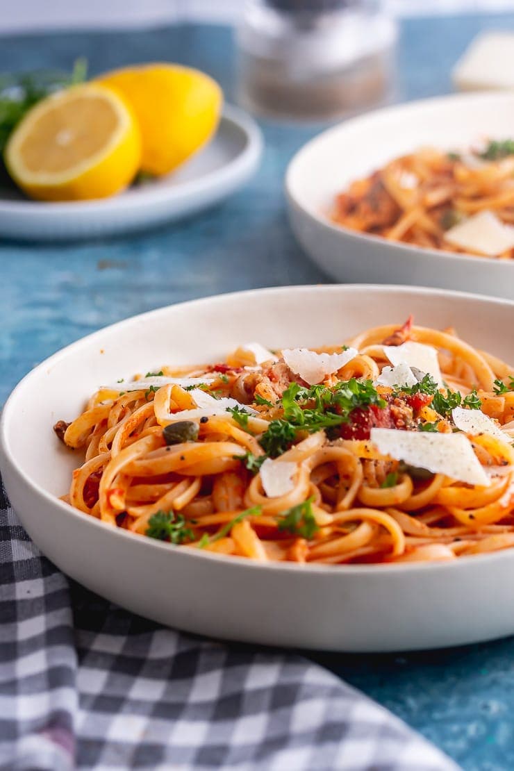 Side on shot of spicy salmon linguine in a white bowl on a blue background with lemons in the background