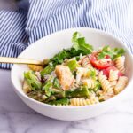 White bowl of mackerel pasta salad on a marble background with a gold fork