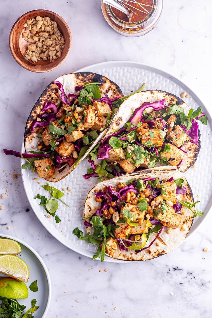 Overhead shot of peanut tofu tacos on a plate with a wooden bowl of chopped peanuts