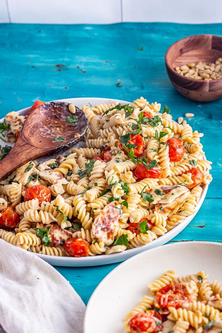 Wooden spoon with halloumi pasta in front of a wooden bowl of pine nuts