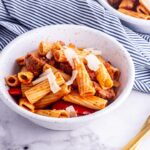 White bowl of sausage pasta with a striped cloth on a marble surface