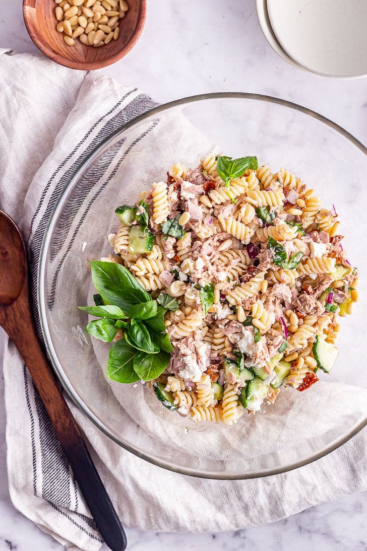 Overhead shot of tuna pasta salad in a glass bowl with a cloth and wooden spoon