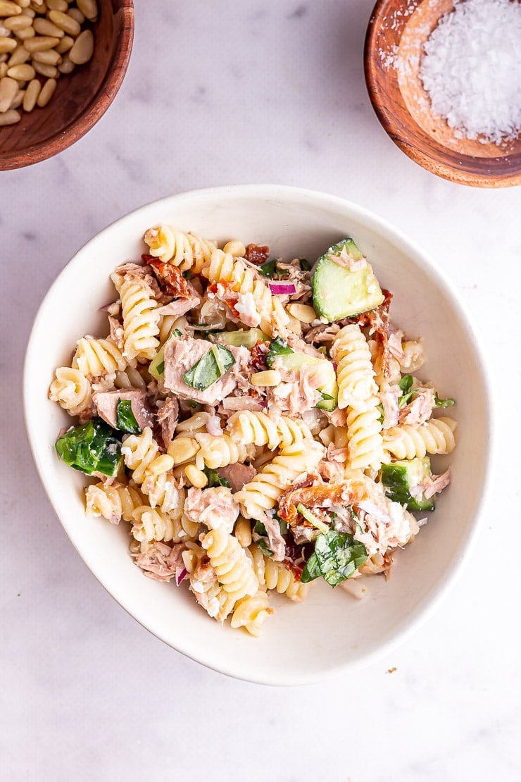 Overhead shot of a white bowl of tuna pasta salad with wooden bowls of salt and pine nuts