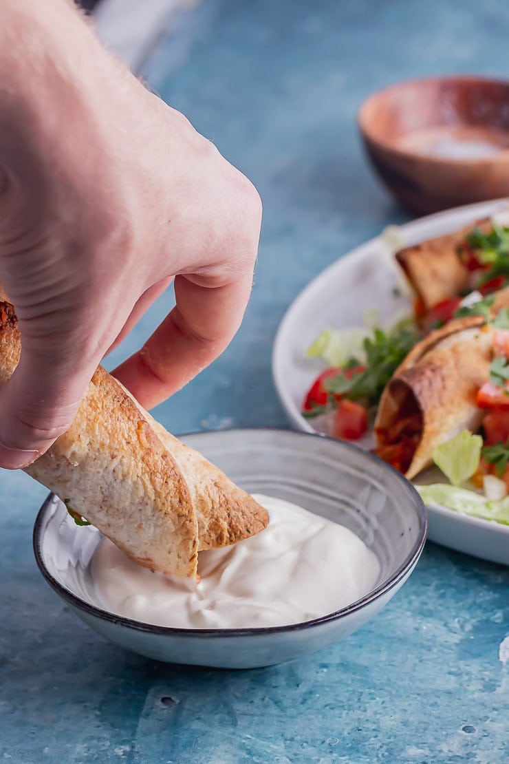 Chicken taquito being dipped in sour cream in a blue bowl