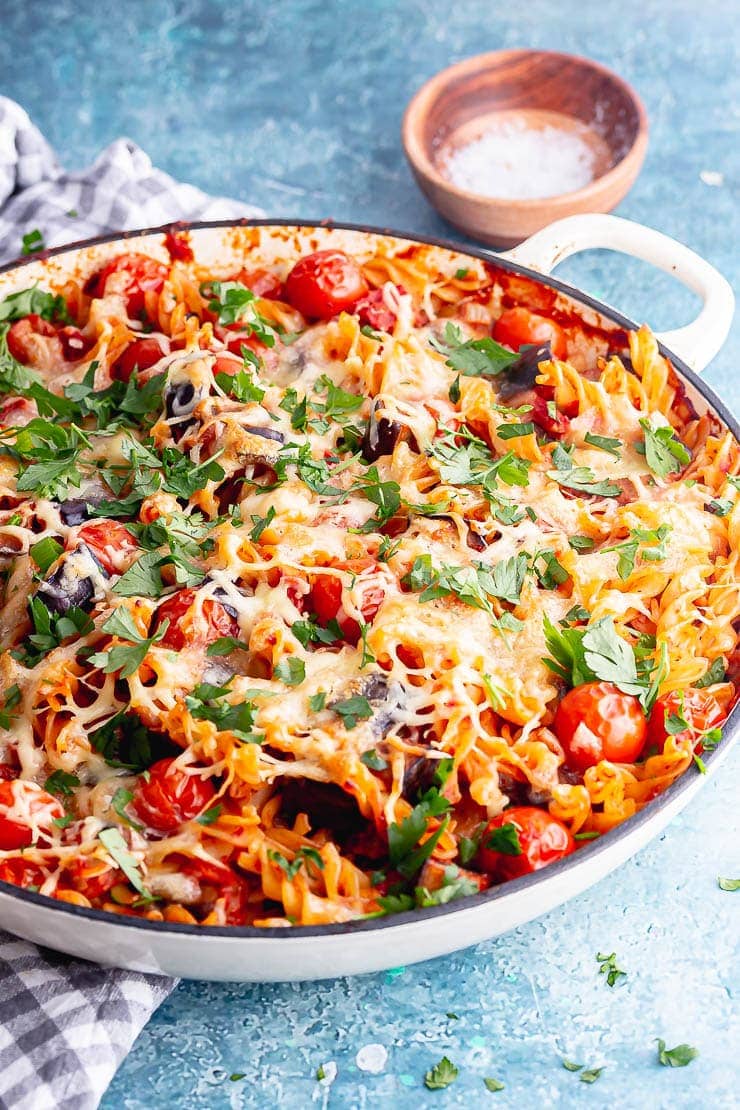 White dish of eggplant baked pasta on a blue background with a wooden bowl of salt in the background