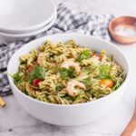 White bowl of pesto pasta salad on a marble background with a wooden spoon