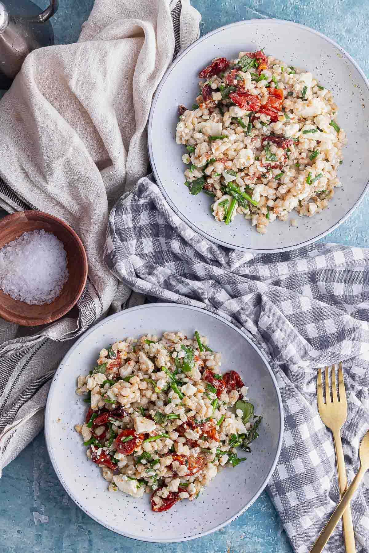 Flat lay shot of two bowls of barley salad with cloths and a wooden bowl of salt
