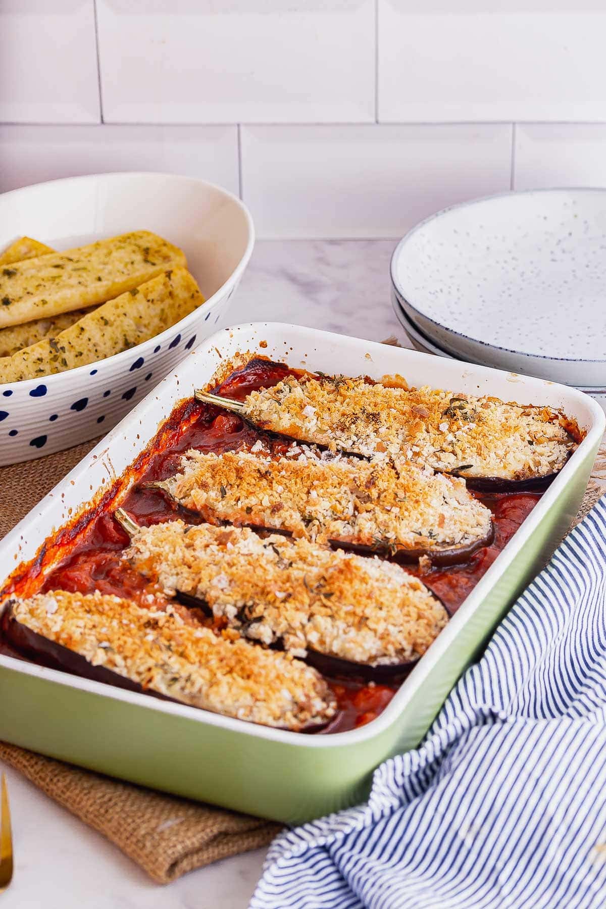 Green baking dish of baked aubergine in tomato sauce with a striped cloth